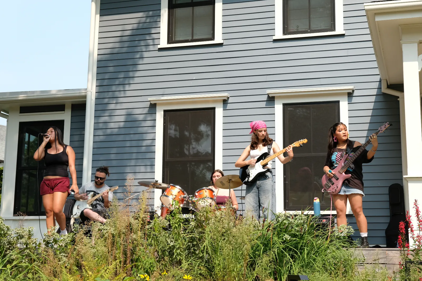 Full band with vocalist, guitars, drums performing on porch wide shot